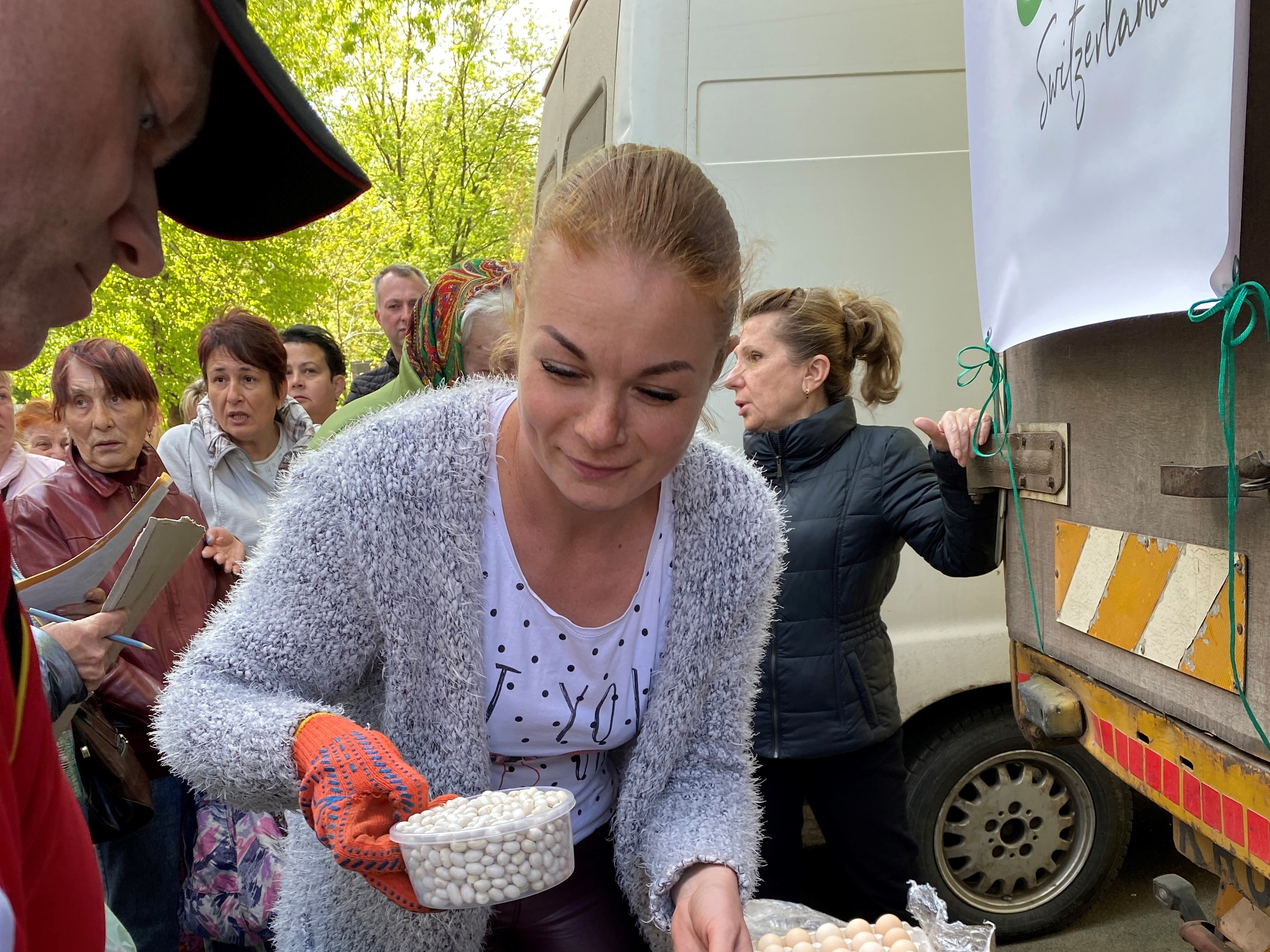 A woman distributes beans and eggs to victims. In the background, several people stand agitated in front of the Green Cross Switzerland food truck.